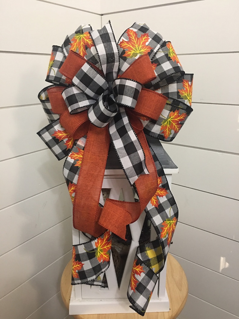 May include: A black and white buffalo plaid ribbon with orange and yellow fall leaves printed on it is tied into a bow on a white lantern.
