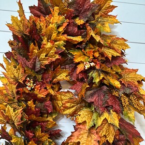 May include: A fall-themed wreath composed of colorful maple leaves in shades of red, orange, and yellow. The wreath is adorned with small, round, berry-like accents, creating a festive and seasonal decoration.