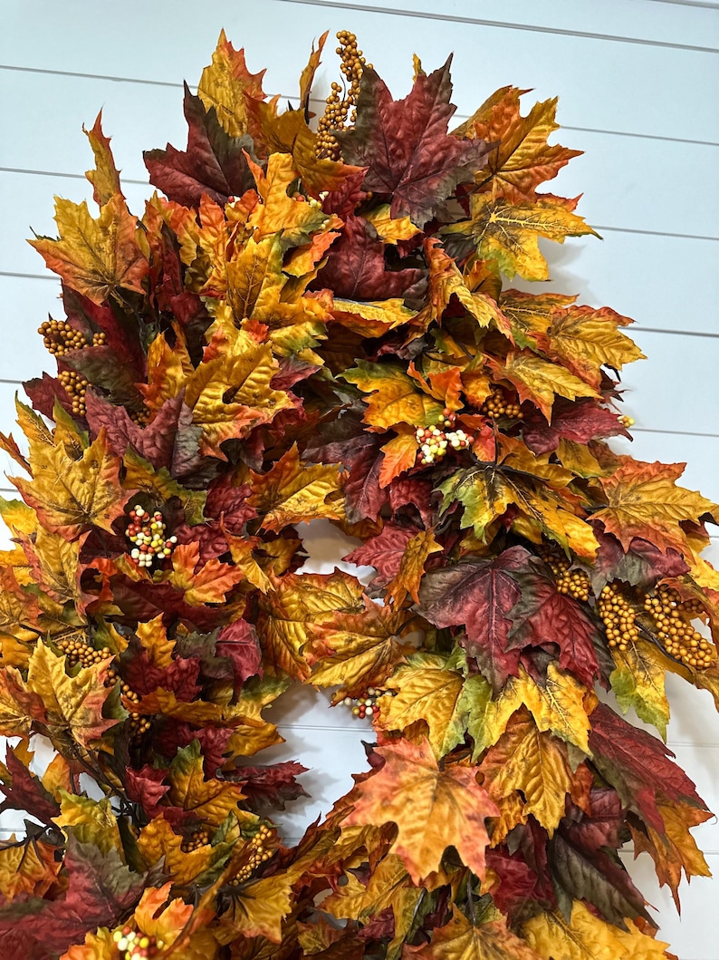 May include: A fall-themed wreath composed of colorful maple leaves in shades of red, orange, and yellow. The wreath is adorned with small, round, berry-like accents, creating a festive and seasonal decoration.