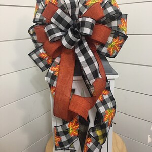 May include: A black and white buffalo plaid ribbon with orange burlap ribbon and a fall leaf print. The ribbon is tied into a bow and is attached to a white lantern.