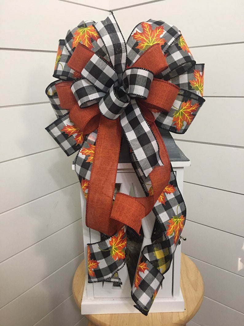 May include: A black and white buffalo plaid ribbon with orange burlap ribbon and a fall leaf print. The ribbon is tied into a bow and is attached to a white lantern.