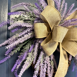 May include: A lavender wreath with a burlap bow. The wreath is made of artificial lavender sprigs and is decorated with a large, rustic burlap bow.