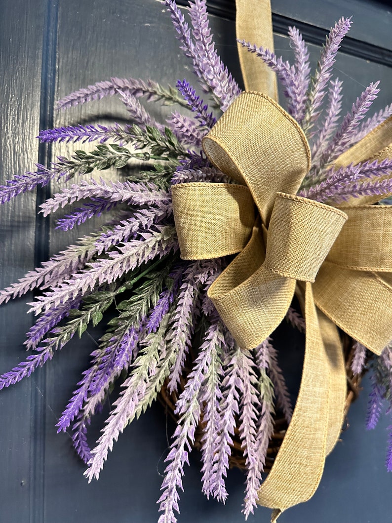 May include: A lavender wreath with a burlap bow. The wreath is made of artificial lavender sprigs and is decorated with a large, rustic burlap bow.