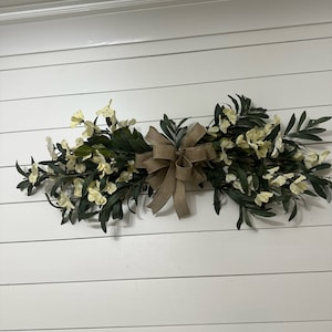 May include: A white and green floral swag with a brown burlap bow, hanging on a white shiplap wall.