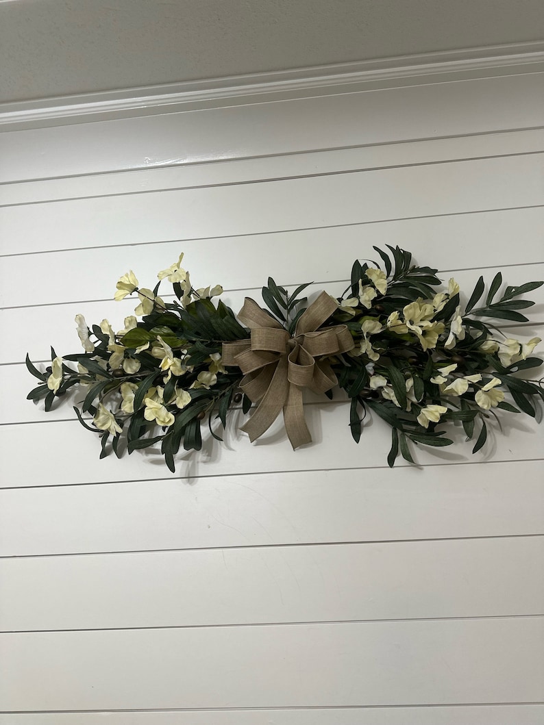 May include: A white and green floral swag with a brown burlap bow, hanging on a white shiplap wall.