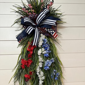 May include: A patriotic floral arrangement featuring red, white, and blue accents. The design includes a large navy blue bow with white stripes, red and white striped ribbons, and faux flowers in red, white, and blue. Green foliage completes the design.