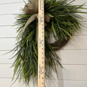 May include: A decorative wreath featuring faux green foliage and a brown burlap bow. The wreath is approximately 28 inches long, with a natural twig base. The greenery has a realistic appearance, suitable for seasonal home decor.
