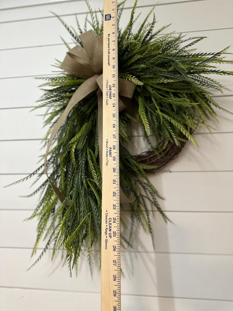 May include: A decorative wreath featuring faux green foliage and a brown burlap bow. The wreath is approximately 28 inches long, with a natural twig base. The greenery has a realistic appearance, suitable for seasonal home decor.