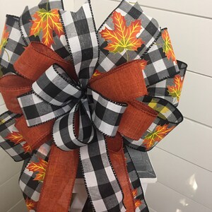 May include: A large, decorative bow made with black and white buffalo check ribbon, orange burlap ribbon, and a ribbon with a fall leaf print. The bow is tied with a loop and tails.