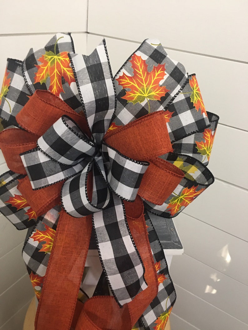 May include: A large, decorative bow made with black and white buffalo check ribbon, orange burlap ribbon, and a ribbon with a fall leaf print. The bow is tied with a loop and tails.