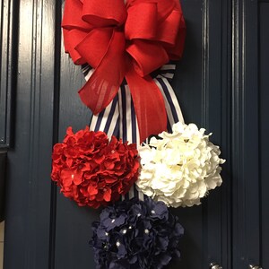 May include: A patriotic-themed decorative accent featuring a large red bow with a blue and white striped ribbon. Below the bow are three floral spheres in red, white, and navy blue, attached to a dark blue cabinet door.