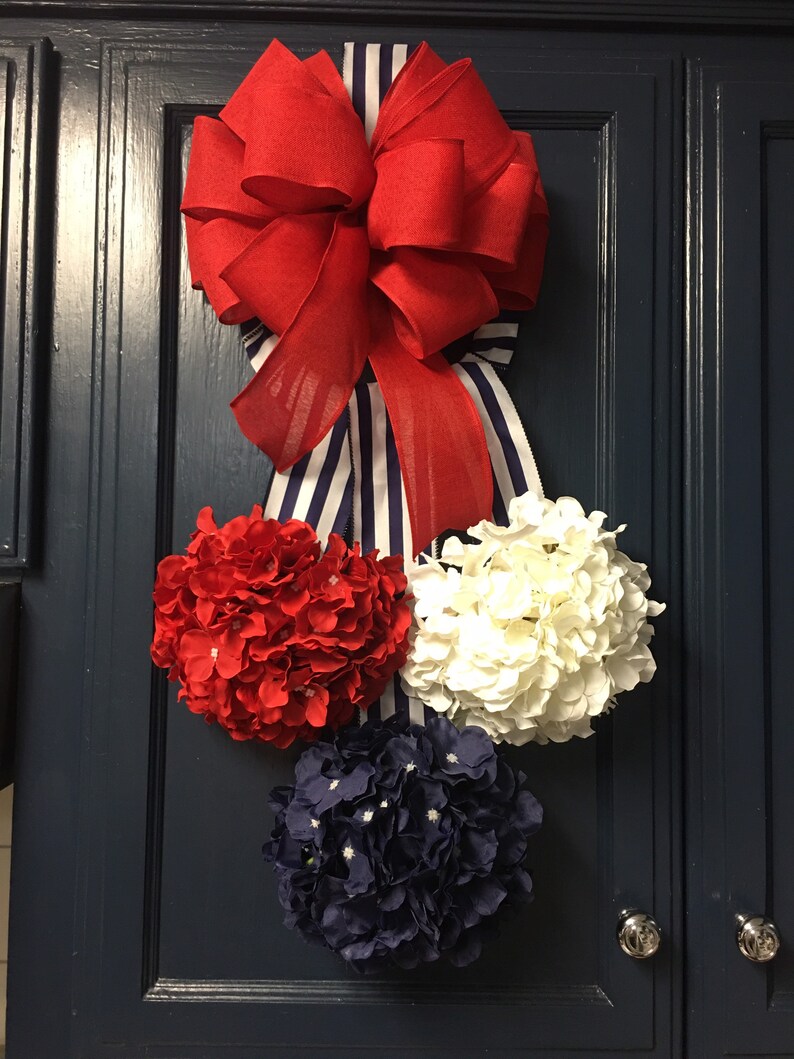 May include: A patriotic-themed decorative accent featuring a large red bow with a blue and white striped ribbon. Below the bow are three floral spheres in red, white, and navy blue, attached to a dark blue cabinet door.