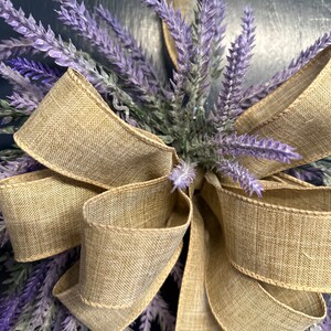 May include: A lavender wreath with a large, rustic brown burlap bow. The wreath is made of artificial lavender sprigs and is perfect for a farmhouse or country-style decor.