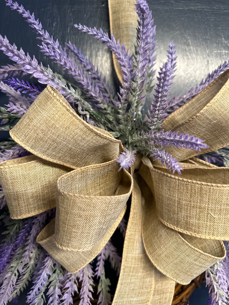 May include: A lavender wreath with a large, rustic brown burlap bow. The wreath is made of artificial lavender sprigs and is perfect for a farmhouse or country-style decor.