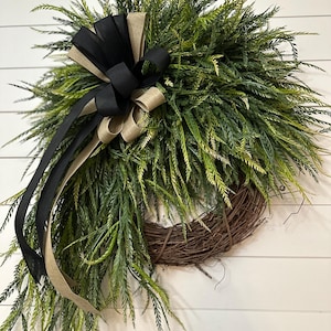 May include: A decorative wreath featuring a natural twig base, lush green foliage, and a large bow. The bow is made of black and tan ribbons. This wreath is suitable for indoor or outdoor use.