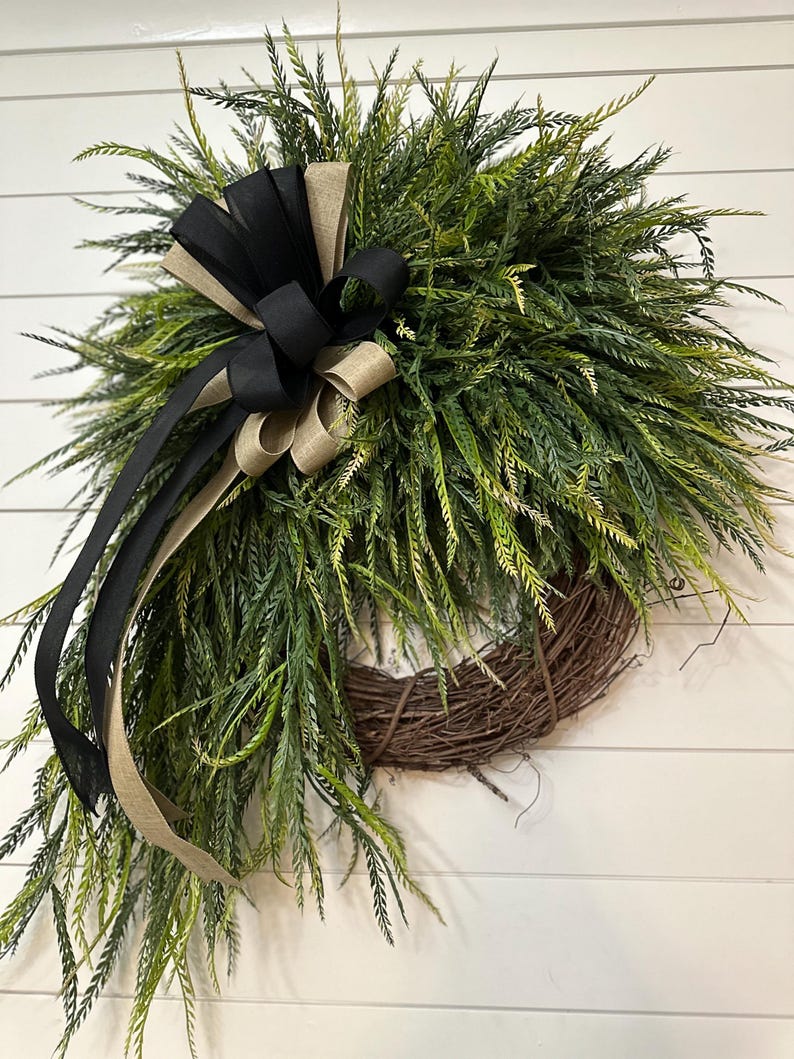 May include: A decorative wreath featuring a natural twig base, lush green foliage, and a large bow. The bow is made of black and tan ribbons. This wreath is suitable for indoor or outdoor use.