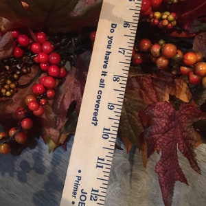 May include: A fall-themed wreath with red berries, orange berries, and brown leaves. A wooden ruler is placed diagonally across the wreath, measuring approximately 10 inches. The ruler has the text "Do you have it all covered?"