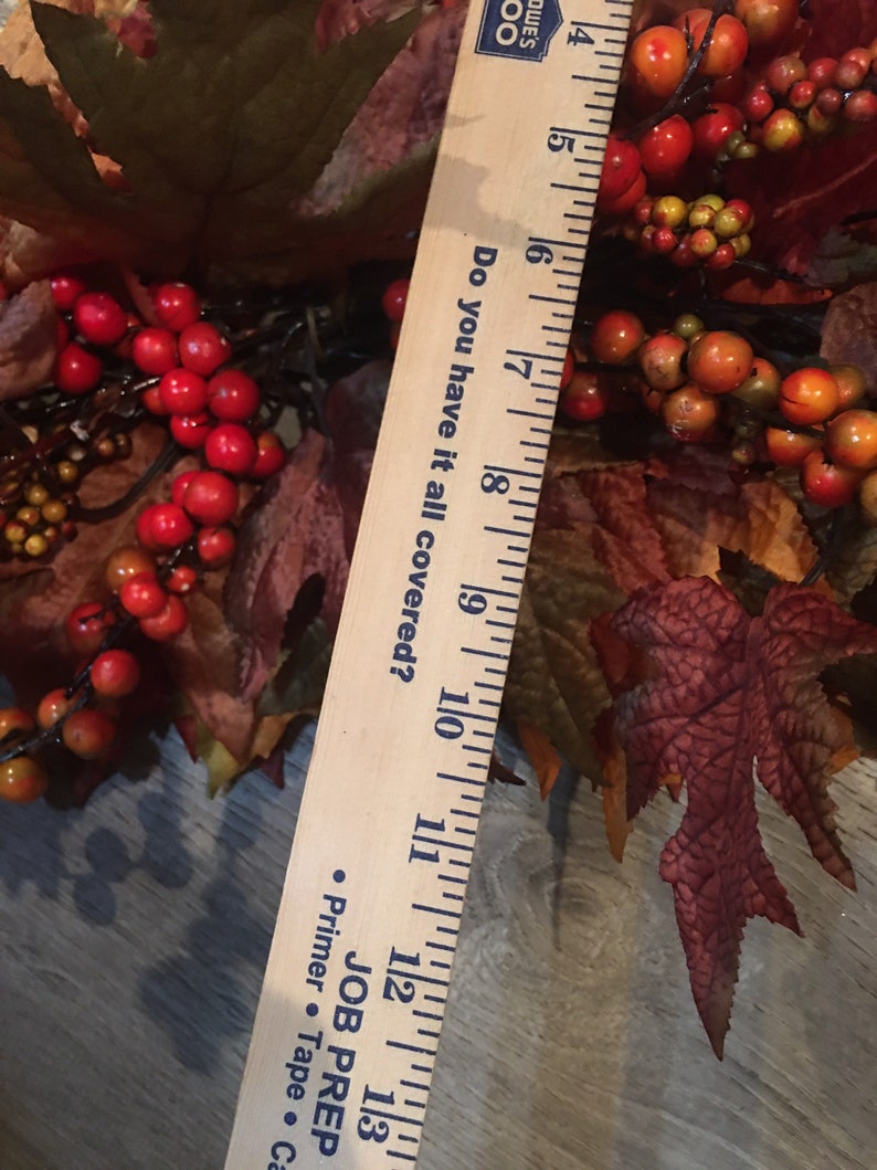 May include: A fall-themed wreath with red berries, orange berries, and brown leaves. A wooden ruler is placed diagonally across the wreath, measuring approximately 10 inches. The ruler has the text "Do you have it all covered?"