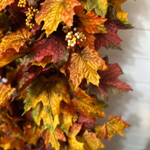May include: A close-up of a fall-themed garland featuring maple leaves in shades of yellow, orange, and red. Small, round, yellow and orange berries are interspersed among the leaves. The leaves have a realistic texture and are arranged to create a full, decorative display.