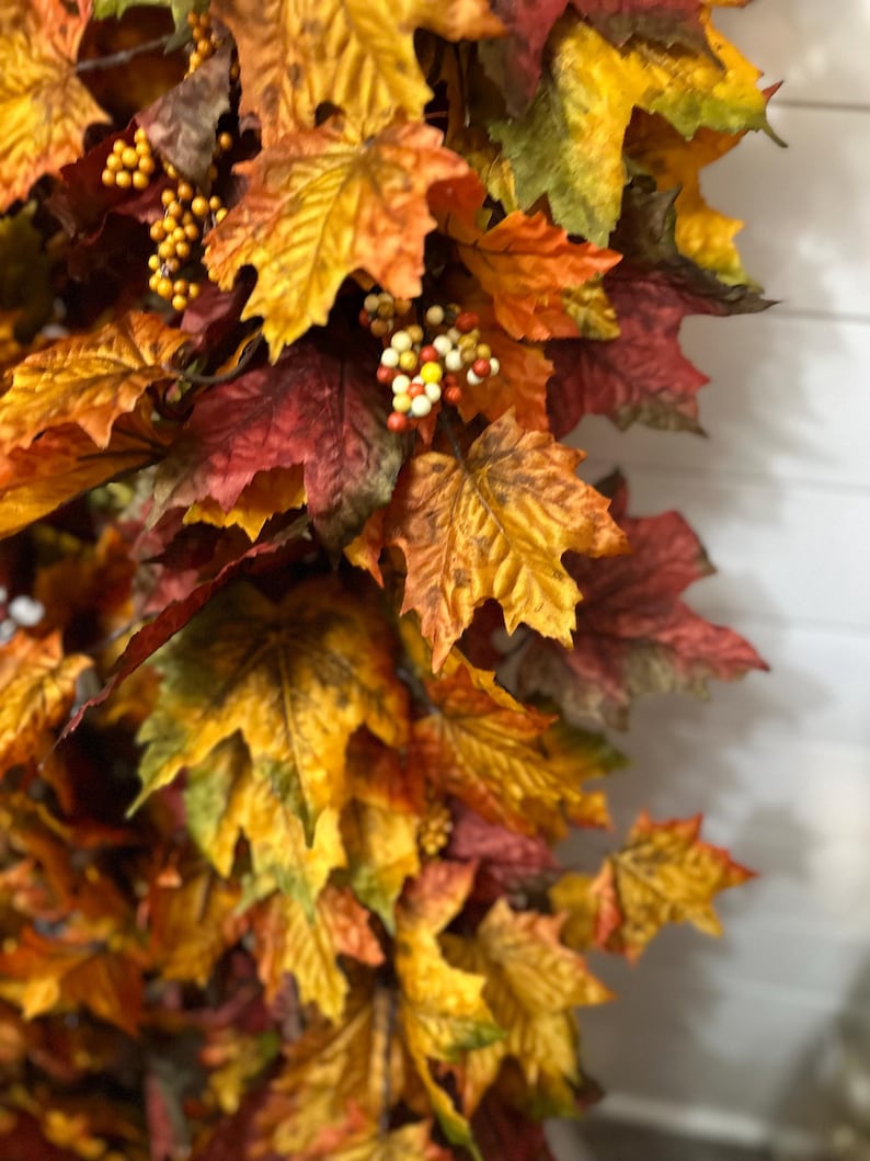 May include: A close-up of a fall-themed garland featuring maple leaves in shades of yellow, orange, and red. Small, round, yellow and orange berries are interspersed among the leaves. The leaves have a realistic texture and are arranged to create a full, decorative display.
