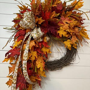 May include: A decorative wreath featuring a grapevine base adorned with artificial autumn leaves in shades of red, orange, and yellow. A cream-colored ribbon with a gold pumpkin print is tied in a bow, with long tails cascading down the side.