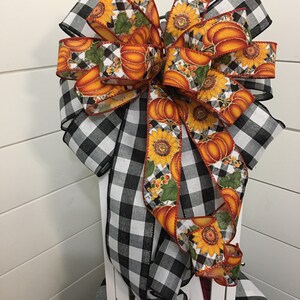 May include: A large, black and white buffalo plaid bow with a pumpkin and sunflower ribbon. The bow is tied around a white lantern.