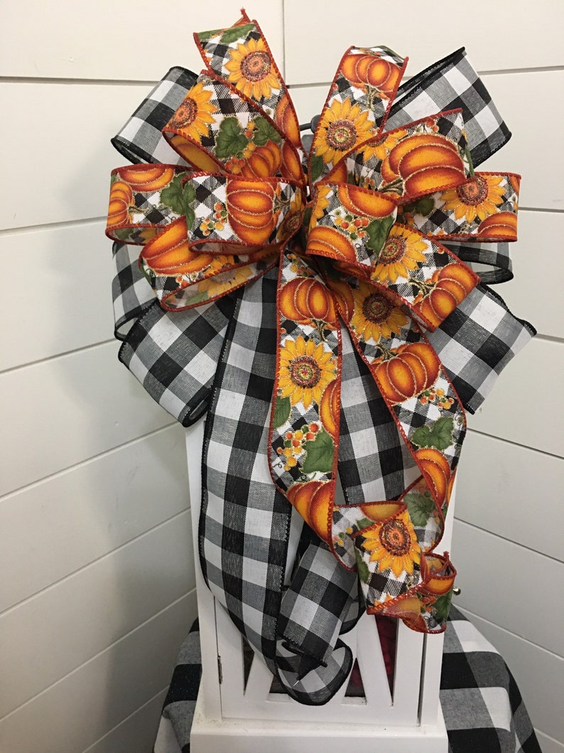 May include: A large, black and white buffalo plaid bow with a pumpkin and sunflower ribbon. The bow is tied around a white lantern.