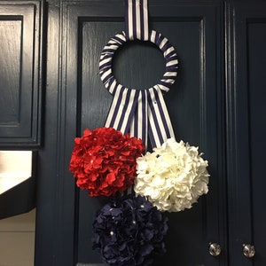 May include: A decorative wreath with a striped ribbon and three floral clusters in red, white, and navy blue. The wreath is hanging on a dark blue cabinet door with silver knobs, adding a patriotic touch to the kitchen decor.