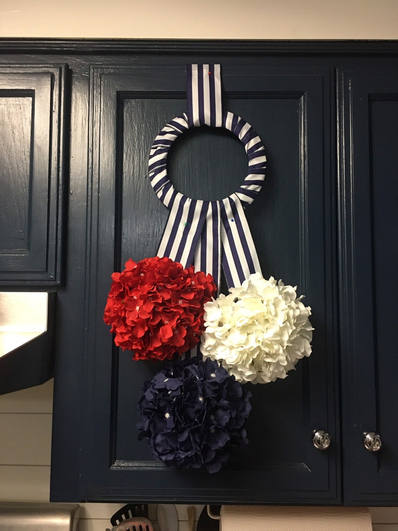 May include: A decorative wreath with a striped ribbon and three floral clusters in red, white, and navy blue. The wreath is hanging on a dark blue cabinet door with silver knobs, adding a patriotic touch to the kitchen decor.