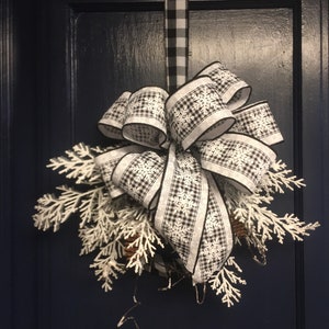 May include: A black and white checkered ribbon bow with snowflake patterns, tied around a white frosted evergreen sprig, hanging on a dark blue door.