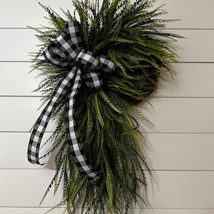 May include: A decorative wreath featuring a cluster of green, needle-like foliage. A large black and white checkered bow is centered on the wreath, with long ribbon tails cascading down. The wreath is mounted on a white, horizontal-slatted background.