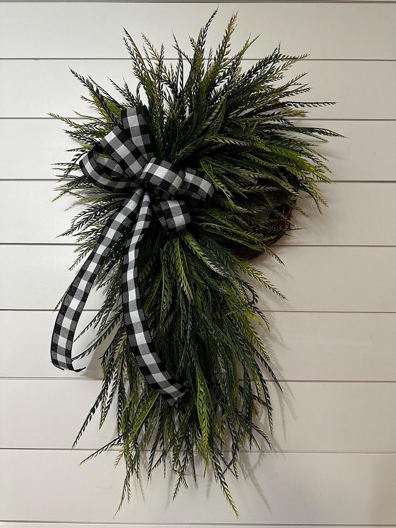 May include: A decorative wreath featuring a cluster of green, needle-like foliage. A large black and white checkered bow is centered on the wreath, with long ribbon tails cascading down. The wreath is mounted on a white, horizontal-slatted background.