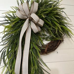 May include: A decorative wreath featuring a circular grapevine base adorned with lush green foliage. A large bow made of striped ribbon in shades of white and tan is centered at the top, with long ribbon tails cascading down. The wreath is designed for home decor.