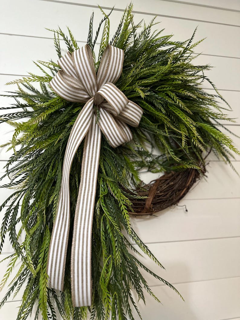 May include: A decorative wreath featuring a circular grapevine base adorned with lush green foliage. A large bow made of striped ribbon in shades of white and tan is centered at the top, with long ribbon tails cascading down. The wreath is designed for home decor.