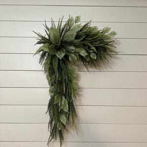 May include: A decorative, artificial greenery arrangement in shades of green. The arrangement features a mix of leafy sprigs and cascading foliage, creating a natural, textured appearance. The piece is designed in a corner shape, suitable for wall decor.
