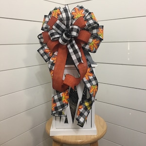 May include: A white lantern with a black and white buffalo plaid ribbon and a ribbon with orange and yellow fall leaves. The lantern is sitting on a wooden stool.