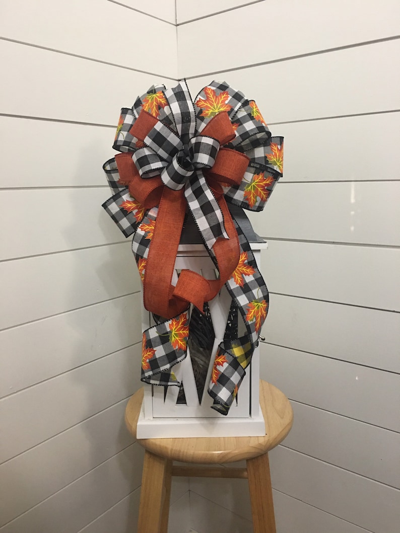 May include: A white lantern with a black and white buffalo plaid ribbon and a ribbon with orange and yellow fall leaves. The lantern is sitting on a wooden stool.