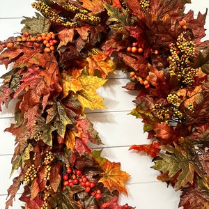 May include: A fall-themed wreath with a variety of colorful leaves in shades of red, orange, and green. The wreath is adorned with clusters of small, yellow and orange berries, creating a festive and seasonal decoration.