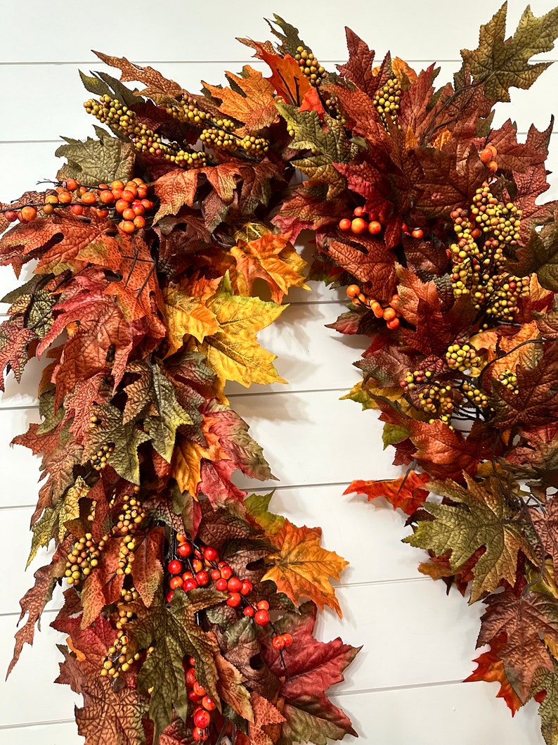 May include: A fall-themed wreath with a variety of colorful leaves in shades of red, orange, and green. The wreath is adorned with clusters of small, yellow and orange berries, creating a festive and seasonal decoration.