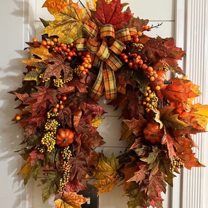 May include: A fall-themed wreath featuring a variety of colorful artificial leaves in shades of orange, red, and yellow. The wreath is adorned with small pumpkins, berries, and a plaid ribbon. This decorative wreath is perfect for autumn home decor.