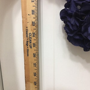 May include: A wooden ruler with black markings, measuring up to 31 inches. The ruler is positioned vertically against a white surface, with a portion of a dark blue floral arrangement visible. Text on the ruler includes "CLEAN UP" and "Cleaner. Rags. Gloves."