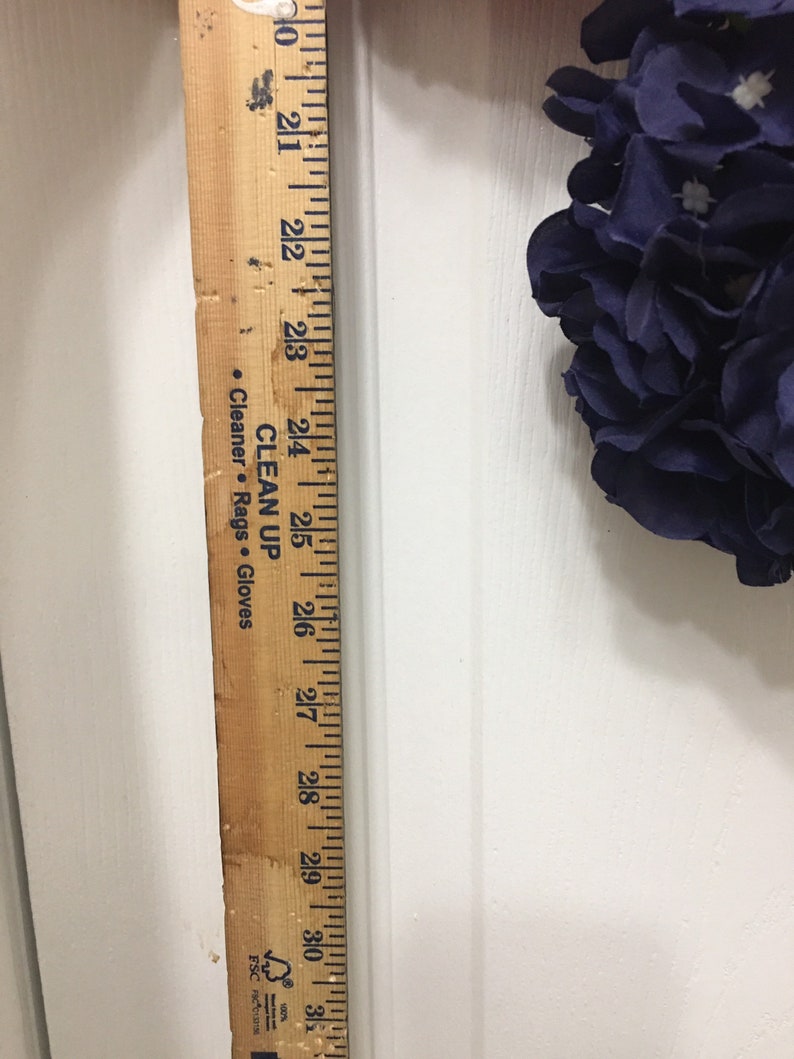 May include: A wooden ruler with black markings, measuring up to 31 inches. The ruler is positioned vertically against a white surface, with a portion of a dark blue floral arrangement visible. Text on the ruler includes "CLEAN UP" and "Cleaner. Rags. Gloves."