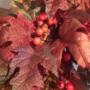 May include: Close-up of a fall-themed decorative arrangement featuring large, textured leaves in shades of red, brown, and orange. Clusters of small, round, bright red berries are interspersed among the leaves, creating a seasonal display.