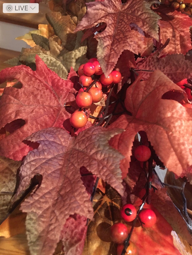 May include: Close-up of a fall-themed decorative arrangement featuring large, textured leaves in shades of red, brown, and orange. Clusters of small, round, bright red berries are interspersed among the leaves, creating a seasonal display.