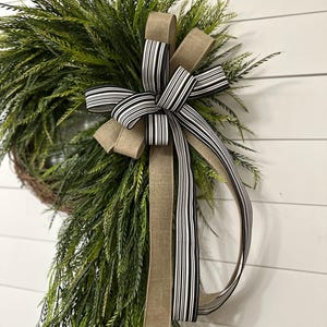 May include: A green wreath with long, thin leaves, accented by a large bow. The bow features a striped black and white ribbon, and a solid tan ribbon. The wreath is displayed against a white, wood-paneled wall.