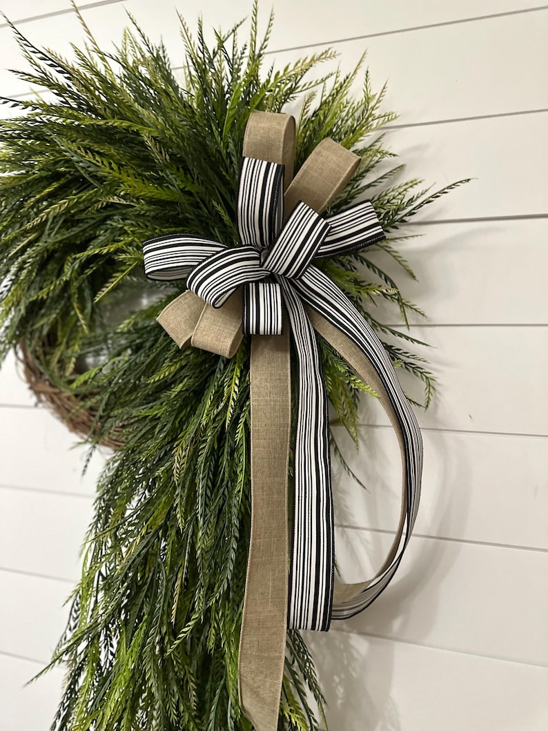May include: A green wreath with long, thin leaves, accented by a large bow. The bow features a striped black and white ribbon, and a solid tan ribbon. The wreath is displayed against a white, wood-paneled wall.