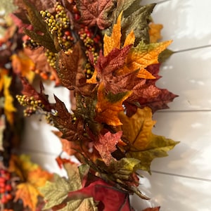 May include: A close-up of a fall-themed wreath with colorful leaves and berries. The leaves are in shades of red, orange, and green, with small yellow berries. The wreath is against a white background.