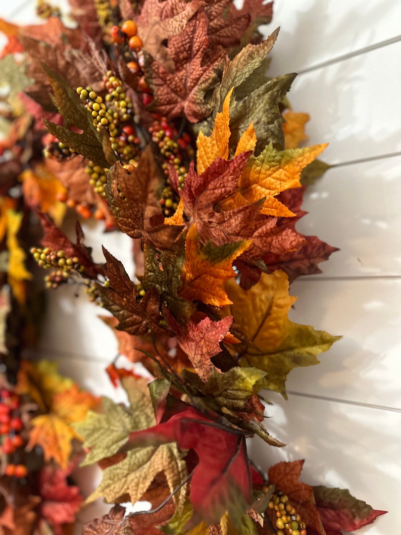 May include: A close-up of a fall-themed wreath with colorful leaves and berries. The leaves are in shades of red, orange, and green, with small yellow berries. The wreath is against a white background.