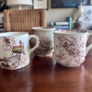 May include: Three antique ceramic mugs with unique designs. One mug features a botanical print with a clock, another has a landscape scene, and the third displays a sailboat and floral pattern in burgundy. The mugs are on a wooden table.