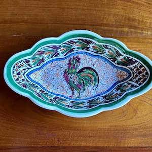 May include: An oval ceramic dish with a scalloped edge, featuring a hand-painted rooster in the centre surrounded by a detailed floral pattern. The rim is decorated with green, brown, and orange leaf motifs on a white background with blue accents.